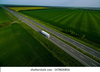 White Truck Driving On Asphalt Road Along The Green Fields. Seen From The Air. Aerial View Landscape. Drone Photography.  Cargo Delivery