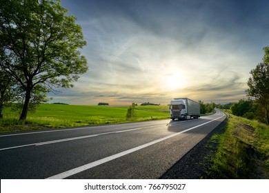 White Truck Driving On The Asphalt Road Next To The Green Field In Rural Landscape At Sunset