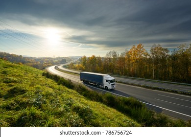 White Truck Driving On The Highway Turning Towards The Horizon In An Autumn Landscape With Sun Shining Through The Clouds In The Sky