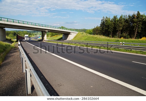 White truck entering under a concrete bridge over a\
highway in a wooded landscape. Noise protection wall. White clouds\
in the blue sky.