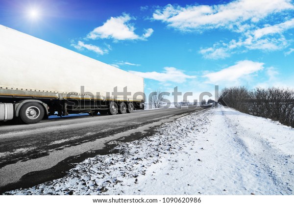 White truck and a white trailer with space for text\
on the winter countryside road with snow against blue sky with\
clouds and bright sun
