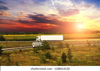 White Truck With A White Trailer And A Van On The Countryside Road With Fields And Forest Against Night Sky With Sunset