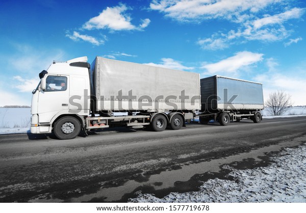 White truck and two trailers with space for text\
on the winter countryside road with snow against blue sky with\
clouds