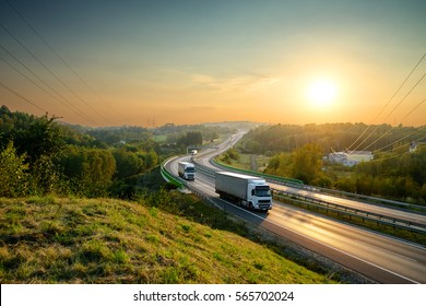 White trucks driving on the highway winding through forested landscape at sunset.