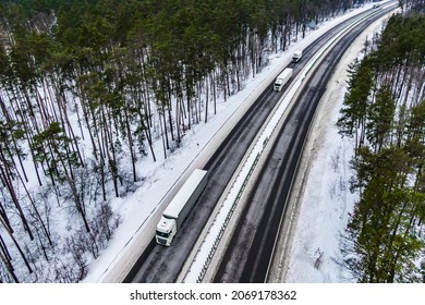 White Trucks On The Higthway. Convoy  Whith Cargo Driving By Road Seen From The Air. Top View Landscape. Shooting From A Drone. Cargo Delivery In Winter. Aerial View Of Snow Road In Winter Forest 