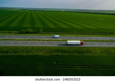 White Trucks On The Higthway Sunset. Cargo Delivery Driving On Asphalt Road Along The Green Fields. Seen From The Air. Aerial View Landscape. Drone Photography.