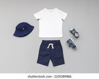 White T-shirt, Blue Shorts, Sandals And Panama Hat On Grey Background. Children's Clothing. Summer Outfit For A Boy. Flat Lay, Top View.