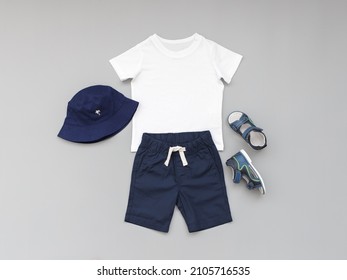 White T-shirt, Blue Shorts, Sandals And Panama Hat On Grey Background. Children's Clothing. Summer Outfit For A Boy. Flat Lay, Top View.