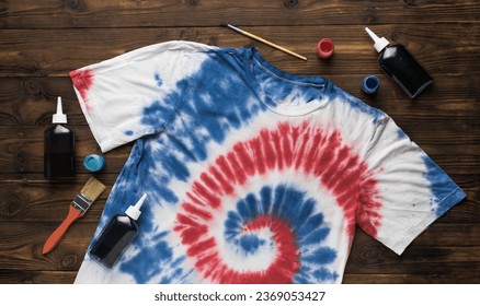 A white T-shirt painted in red and blue tie dye style and materials for coloring on a wooden background. The concept of self-dyeing clothes. Flat lay.