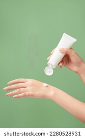 A white tube without label is held in woman model’s hand with a cream texture on another hand. Green pastel background. Branding mockup