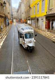 White tuk-tuk vehicle making its way down a narrow, cobblestone street in the historic downtown of Lisbon, Portugal, with tram tracks under its wheels. Selective focus.