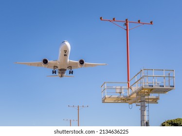 A White Twin Engine Jet Is On Final Approach With The Landing Gear Down.  It Is Low In The Sky And Is Flying Past A Large Tower Of Runway Landing Lights.