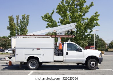 White Utility Truck With Cherry Picker Parked In Parking Lot. Blue Sky And Tree Background. Horizontal.