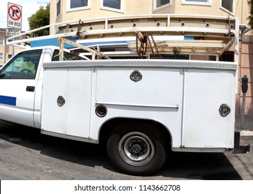 White Utility Truck On Urban Street With Ladders Atop Vehicle..