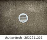 A white vent is on a wall. The vent is round and has a white cap. The vent is on a wall that is tan in color