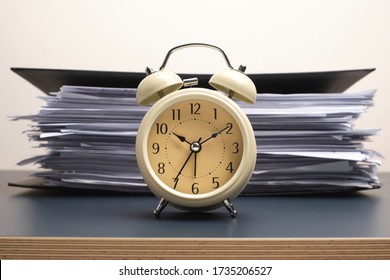 White vintage alarm clock on working desk with many paper files stacked in black folder isolated on white wall background. - Powered by Shutterstock