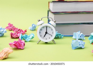 A white vintage clock in the foreground at 7:00 am on a light green background. In the background are books and crumpled colorful paper. Study, school concept. Back to school.