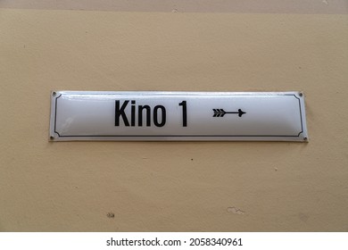 White vintage enamel sign with black inscription "Kino 1" (which means "cinema 1") and a directional arrow pointing to the right. Behind it a beige wall.