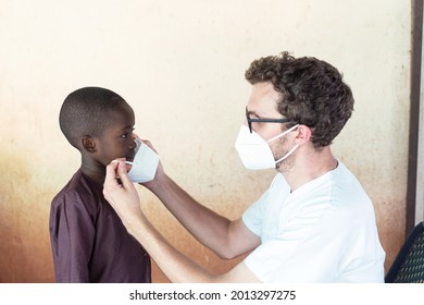 White Volunteering Doctor Helping Boy To Wear His Face Mask To Prevent The Spread Of A Deadly Virus In Africa.