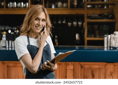 White waitress small business owner bartender barista writing orders in clipboard wearing blue apron talking on phone for takeout takeaway delivery looking at the camera at the bar counter - Powered by Shutterstock