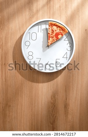 White wall clock with a slice of pizza instead of hands on a light wooden table in soft sunlight. Funny creative Pizza Time concept.