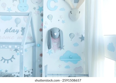 White wall is decorated patterns stars, colorful stickers air ballons, wooden shelf of shape cloud and toy bunny and rabbit in kid room. Stylish interior design child bedroom. Decoration for baby room
