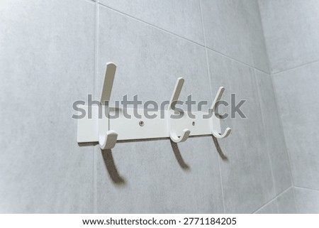 White Wall Hooks Mounted On Grey Tiles, Threeprong Coat Rack With Visible Mounting Screws, Plastic Fixture Suited For Bathroom Or Lockerroom, Minimal Commercial Hardware For Neat