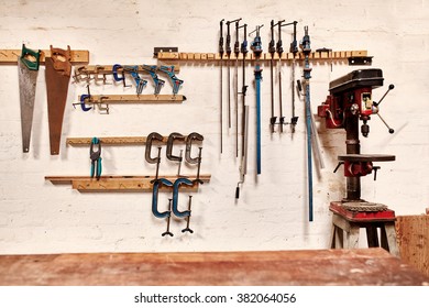 White Wall Of A Woodwork Workshop With Hand Tools Hanging In Neat Rows, A Well Used Drill Press On One Side And A Wooden Work Bench In The Foreground