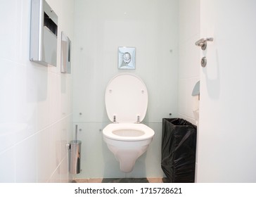 White Water Closet, Restaurant Bathroom, Small Toilet Room With A Wall Mounted, Wall Hung Ceramic Toilet Bowl, Toilet Brush, Waste Basket, Toilet Paper Holder And A Hand Dryer.