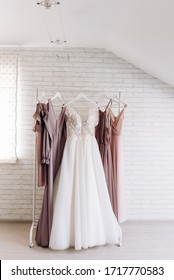 White Wedding Dress And Bridesmaids Dresses Hang On White Velvet Hangers, On The Background Of White Wall With Bricks.