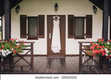 White Wedding Dress Hanging On A Door Of Vintage House With Flowers On Foreground