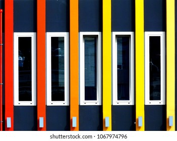 white windows on blue-gray elevation with bright color vertical decorative bands and rectangular light flood light fixtures and strong reflection of the street in the glass. frontal flat on view.  