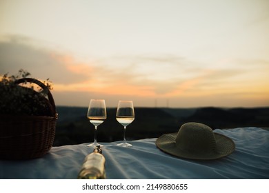 White Wine Bottle, Wine Glasses, Basket With White Flowers And Straw Hat At The Summer Picnic At Sunset.