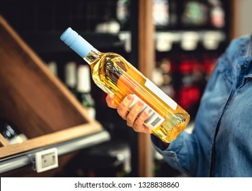 White Wine Bottle In Hand In Liquor Store. Customer Buying Alcohol. Woman Choosing The Right Bottle Of Chardonnay Or Riesling. Alcoholic Beverages Aisle And Shelf In Supermarket. Lady Shopping Drinks.