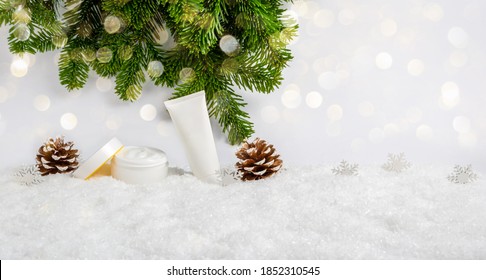 White Winter Skin Care Cosmetic Products In Snow With Christmas Tree, Pine Cones And Bokeh Lights. Opened Face Cream Jar And Hand Creme Or Body Lotion Tube. Festive Banner With Copy Space For Text