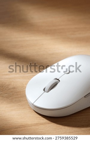 White wireless computer mouse on a light wooden desk surface under soft natural sunlight with soft shadows. Top closeup view with copy space.