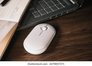 A white wireless mouse and an open notebook with a black pen rest on a dark wood desk next to a modern silver laptop. The scene conveys productivity, technology, and a clean aesthetic.