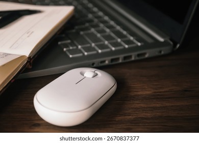 A white wireless mouse and an open notebook with a black pen rest on a dark wood desk next to a modern silver laptop. The scene conveys productivity, technology, and a clean aesthetic.