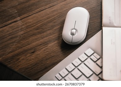 A white wireless mouse and an open notebook with a black pen rest on a dark wood desk next to a modern silver laptop. The scene conveys productivity, technology, and a clean aesthetic.