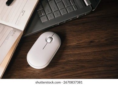 A white wireless mouse and an open notebook with a black pen rest on a dark wood desk next to a modern silver laptop. The scene conveys productivity, technology, and a clean aesthetic.