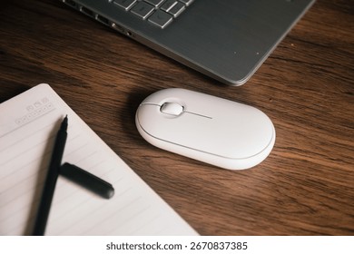A white wireless mouse and an open notebook with a black pen rest on a dark wood desk next to a modern silver laptop. The scene conveys productivity, technology, and a clean aesthetic.