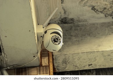 White wireless security camera mounted on the corner of an aged, textured exterior wall. Equipped with signal antenna and infrared LEDs, used for monitoring and securing residential areas.