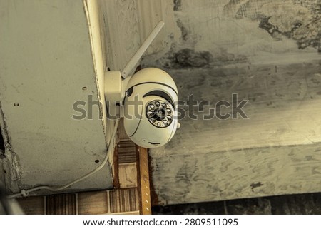 White wireless security camera mounted on the corner of an aged, textured exterior wall. Equipped with signal antenna and infrared LEDs, used for monitoring and securing residential areas.