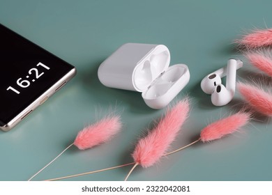 White wireless tws headphones next to a smartphone, a charging case and a pale pink dried flower. Pairing wireless headphones with a smartphone. Concept of relaxing mobile music. Photo. Close-up - Powered by Shutterstock