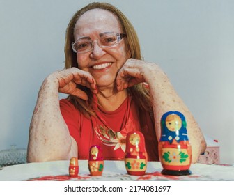 White Woman, Around Seventy Years Old, Long Blond Hair, Wearing Reading Glasses, Wearing A Red T-shirt, Is Smiling And Playing With Matryoshka Dolls. There Is A White Wall In The Background.