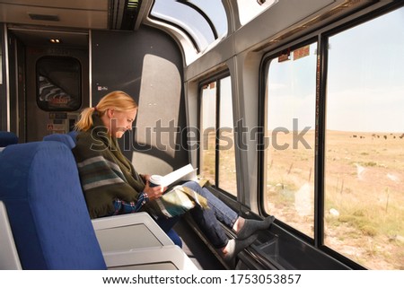 White woman in green scarf around her shoulders sitting in a train lounge room in front of window, reading, drinking coffee. Traveling long distance.