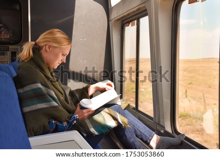 White woman in green scarf around her shoulders sitting in a train lounge room in front of window, reading, drinking coffee. Traveling long distance.