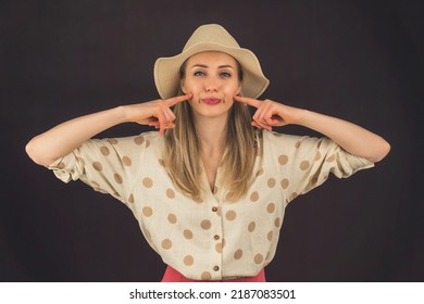 White Woman With Long Blond Hair Wearing Hat And Summery Clothes Putting Pingers To Cheeks Making Silly Expression With Tongue Out. Dark Background Studio Shot. High Quality Photo