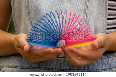 White woman wearing denim overalls playing with the crazy spring toy from the 80s and 90s with predominant yellow, orange, pink and blue colors. The object is held with both hands.