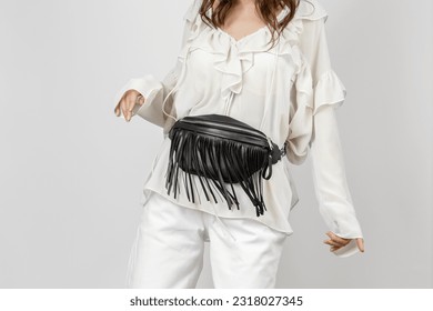 White Woman Wearing White Jeans, Blouse with Frills and Crossbody Black Leather Belt Bag with Decorative Fringe over Grey Background